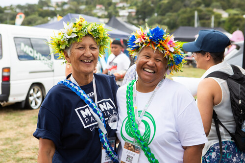 A man and a woman in formal wear stand opposite each other inside a marae, the woman’s arms are stretched out to receive a large intricately woven basket as the man hands it to her, behind the woman, a man in a suit stand solemnly