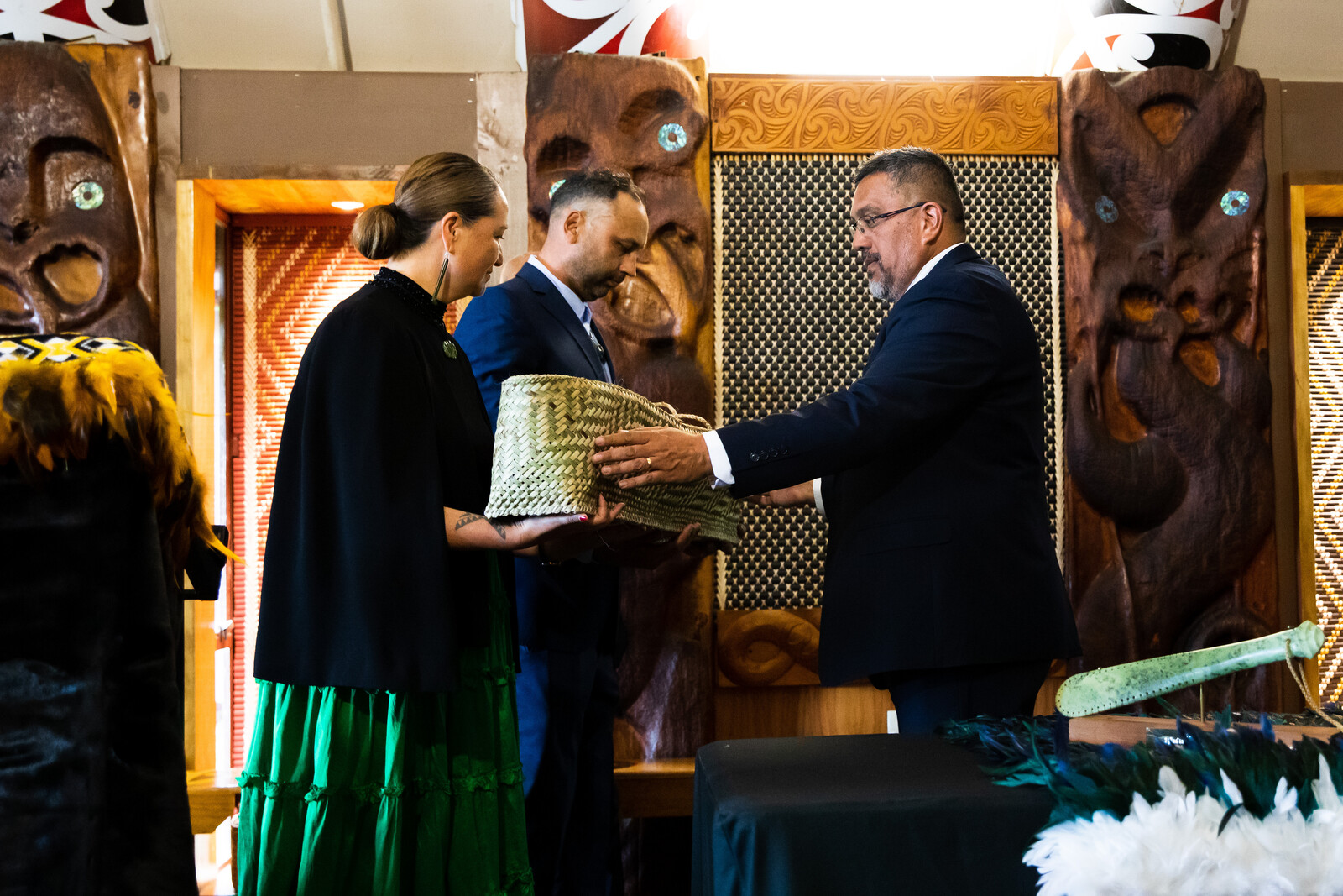 A man and a woman in formal wear stand opposite each other inside a marae, the woman’s arms are stretched out to receive a large intricately woven basket as the man hands it to her, behind the woman, a man in a suit stand solemnly