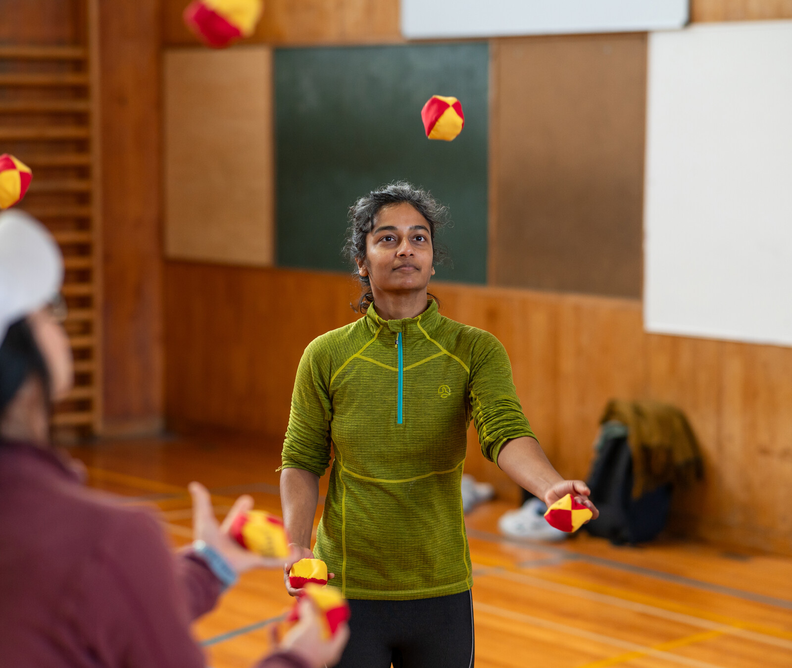 A young woman is juggling red and yellow balls in a gymnasium as another person watches nearby