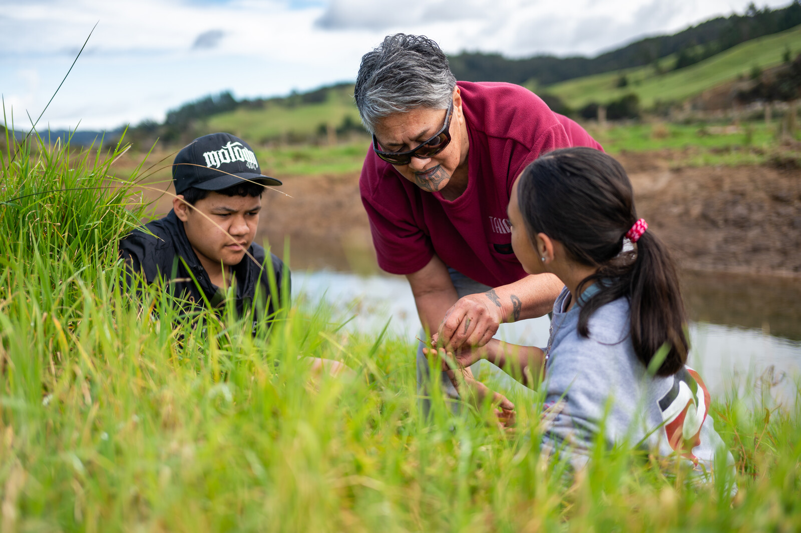 A young boy and girl sit in long grass listening to a woman as she places an unseen object in the young girl's hand