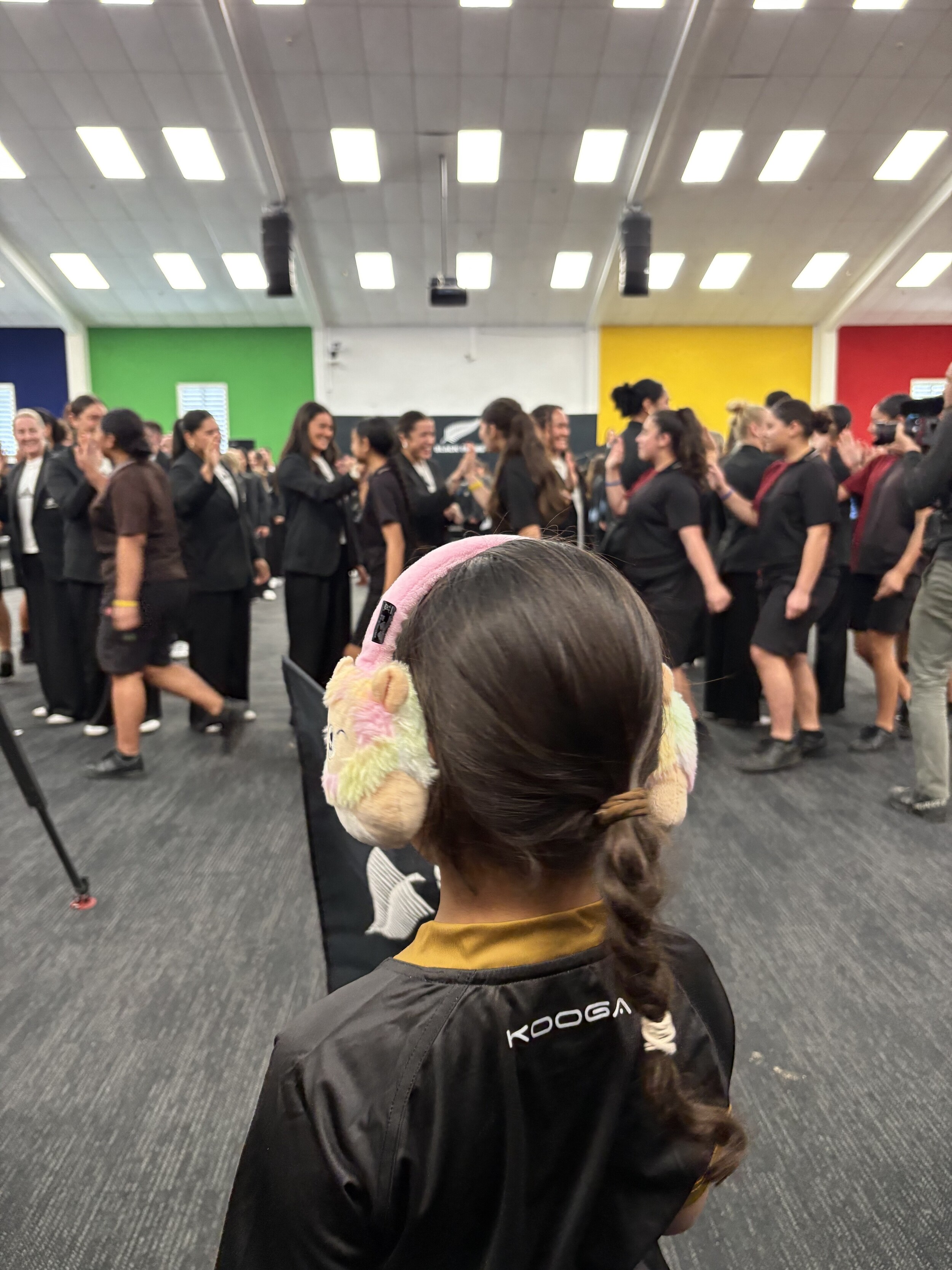 A young person wearing colourful headphones holds a Black Ferns flag while watching a large group of women smiling and hi-fiving each other in the background.
