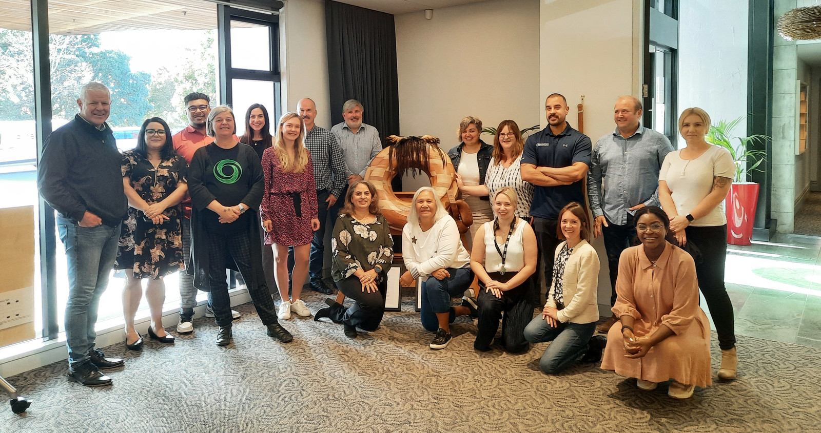 A large group of people stand and crouch around a large, closed koru carved from wood smiling at the camera. 