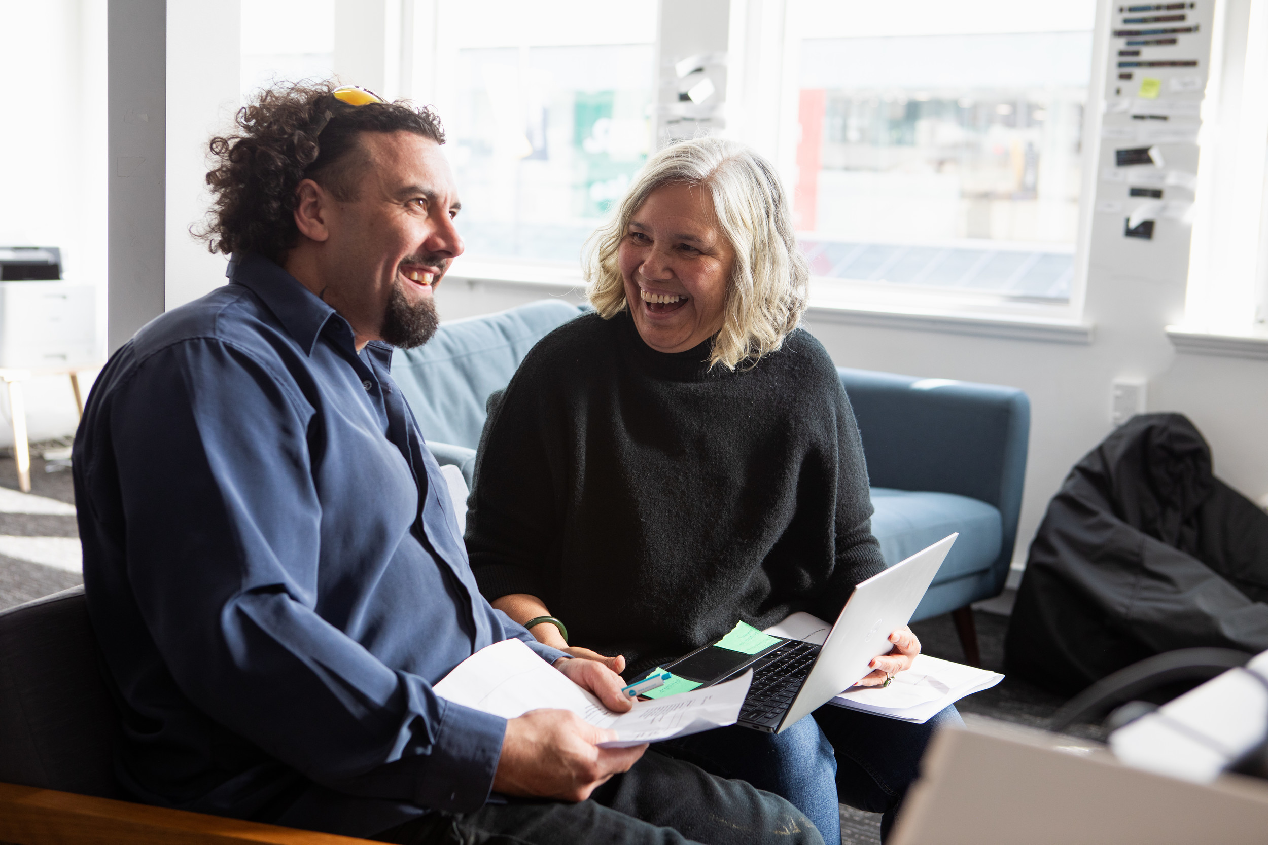 A man and a woman are seated next to each other on a couch, the man holds a piece of paper with both hands, smiling to someone off camera while the woman laughs with the man while holding an open laptop on her lap. 