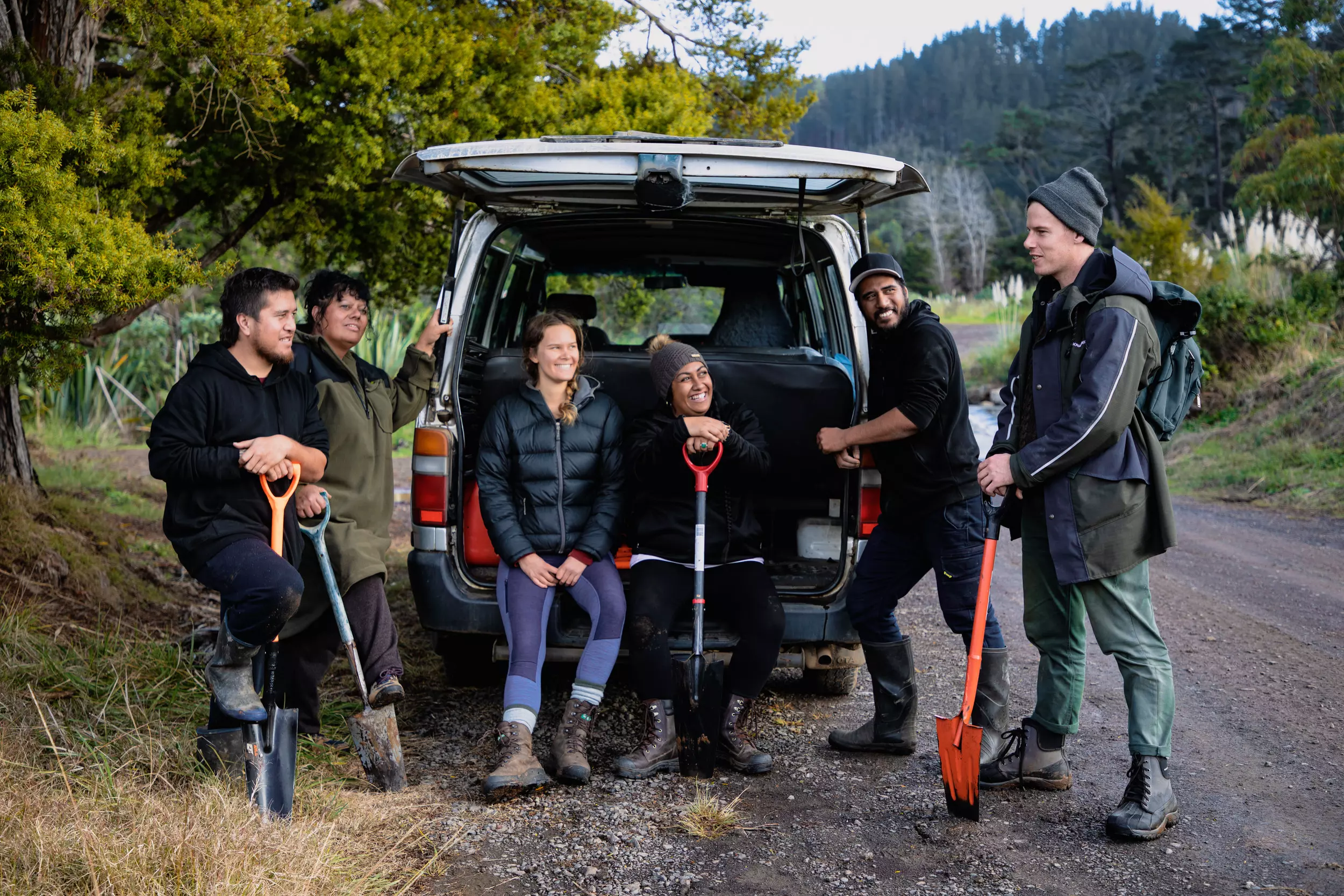 A group of four people in outdoor gear holding large shovels stand around the open boot of a van parked on the side of a bush road. Sitting in the boot of a van are two women smiling at the group.  