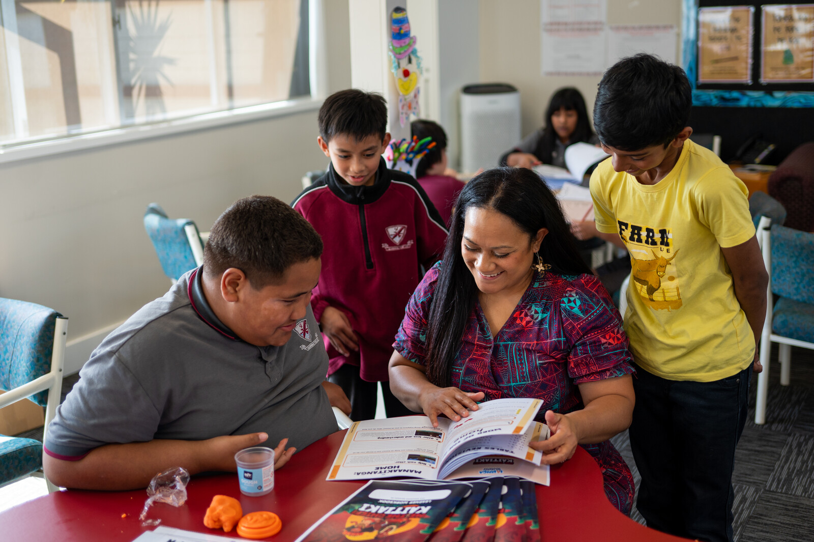A group of three primary students in their uniforms gather around a woman seated at a classroom desk as she reads to them from a workbook