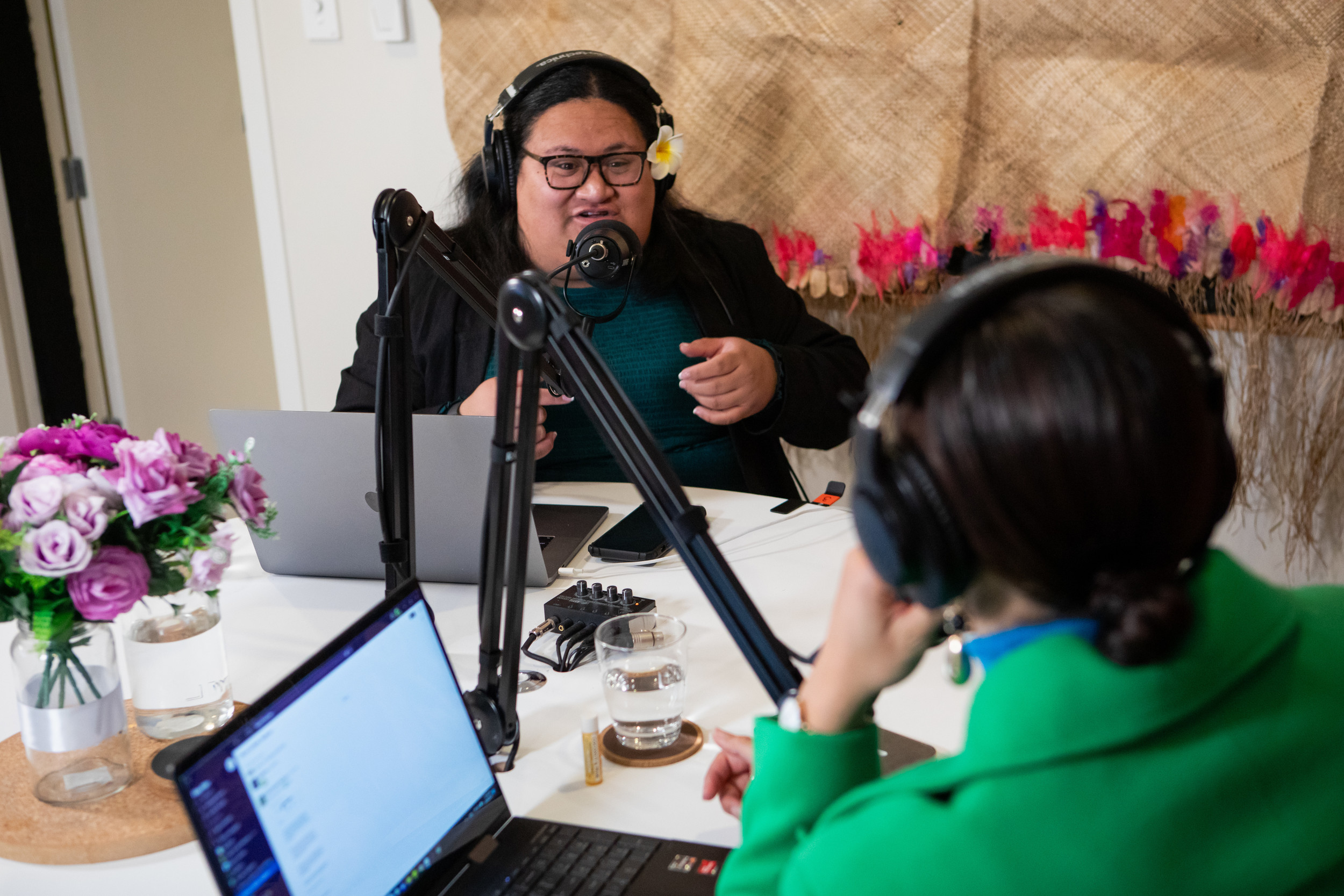 An over the shoulder shot of a woman wearing headphones seated across the table from another woman wearing headphones who is speaking animatedly, both women are speaking into large microphones attached to the table with open laptops in front of them