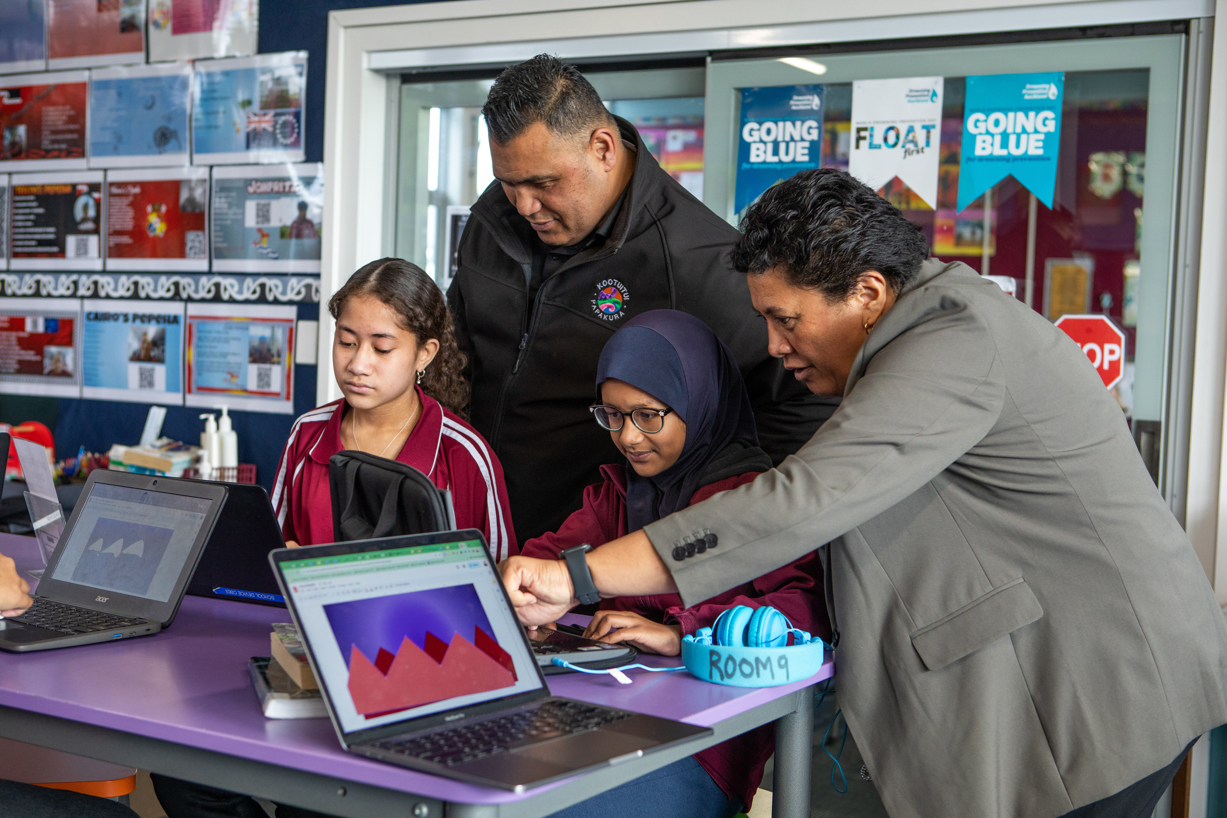 Two primary school girls in a classroom are sitting at a table working on laptops, standing behind them, a man and woman are talking to the girls as the woman points at something on the laptop screen