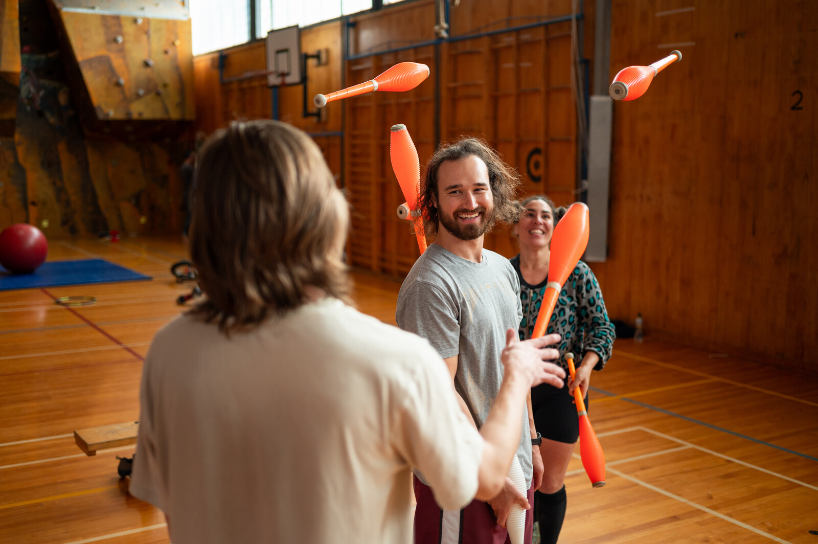 A man smiles at the camera while two people juggle beside him
