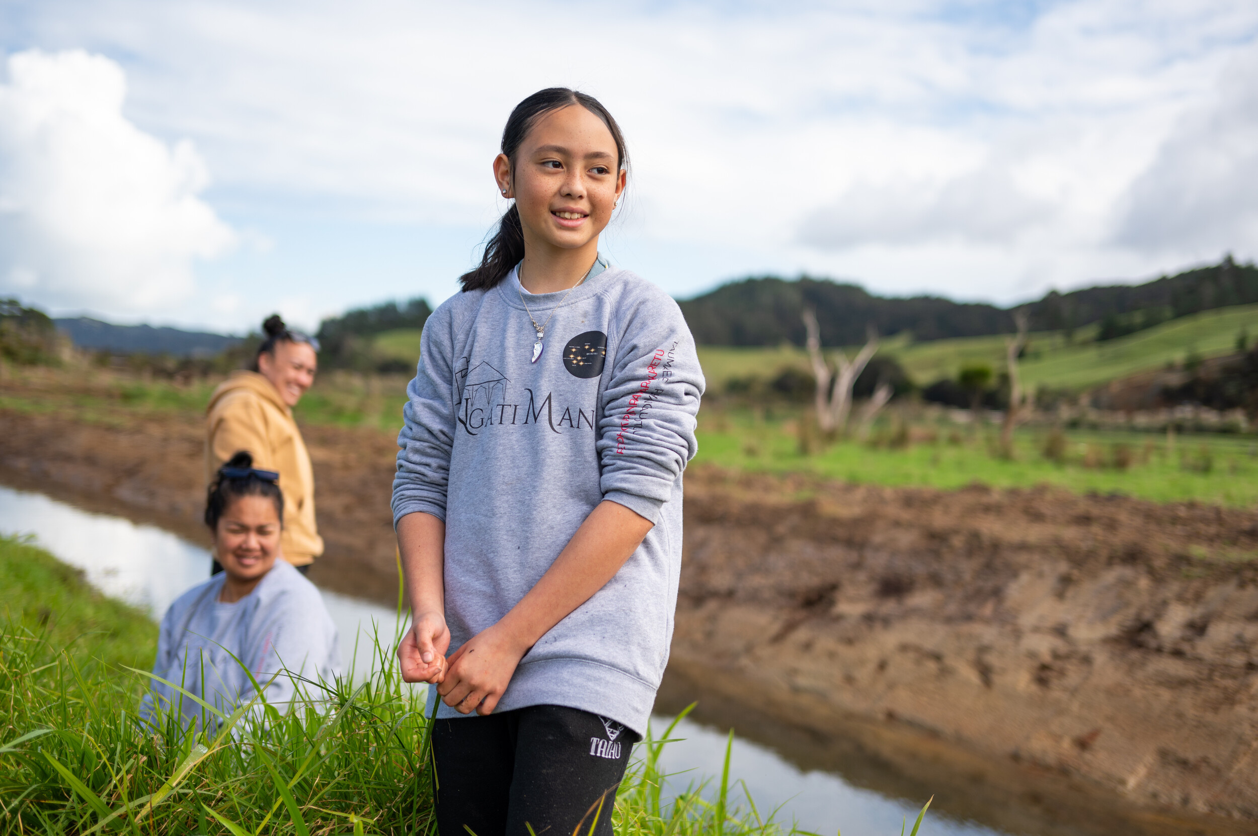 A young girl smiles as she stands in a paddock next to a stream with two women smiling behind her