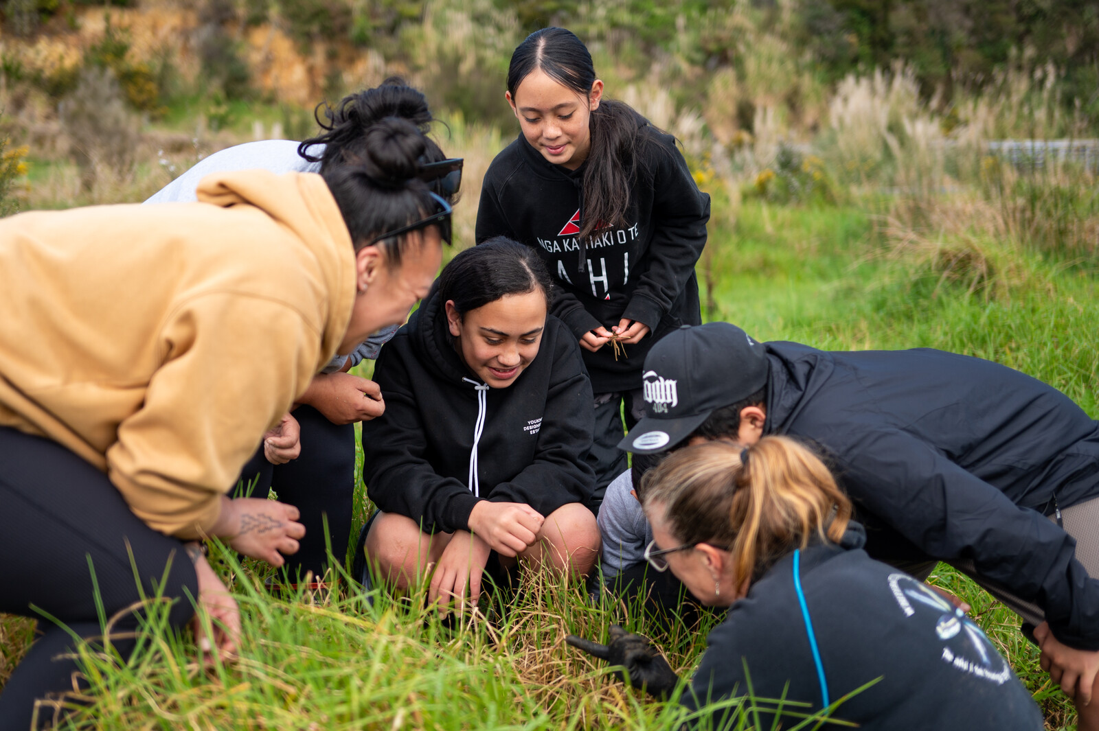2.	A group of young people crouch together outside engrossed in something in the long grass in front of them 