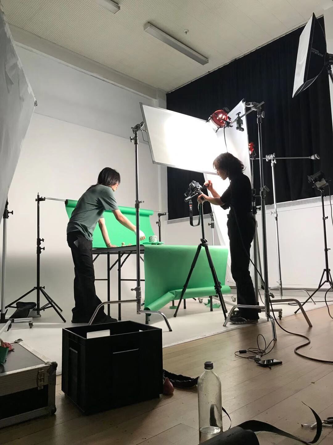 Two men in a photography studio set up lights for a photo shoot with a green cloth as the background.