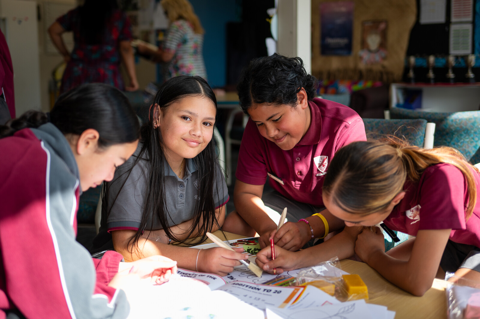 Four smiling primary students are gathered around a table in a classroom reading a workbook together