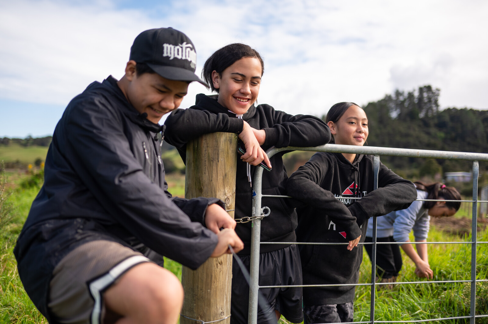 Three young people lean casually on a fence gate smiling under the cloudy sky 