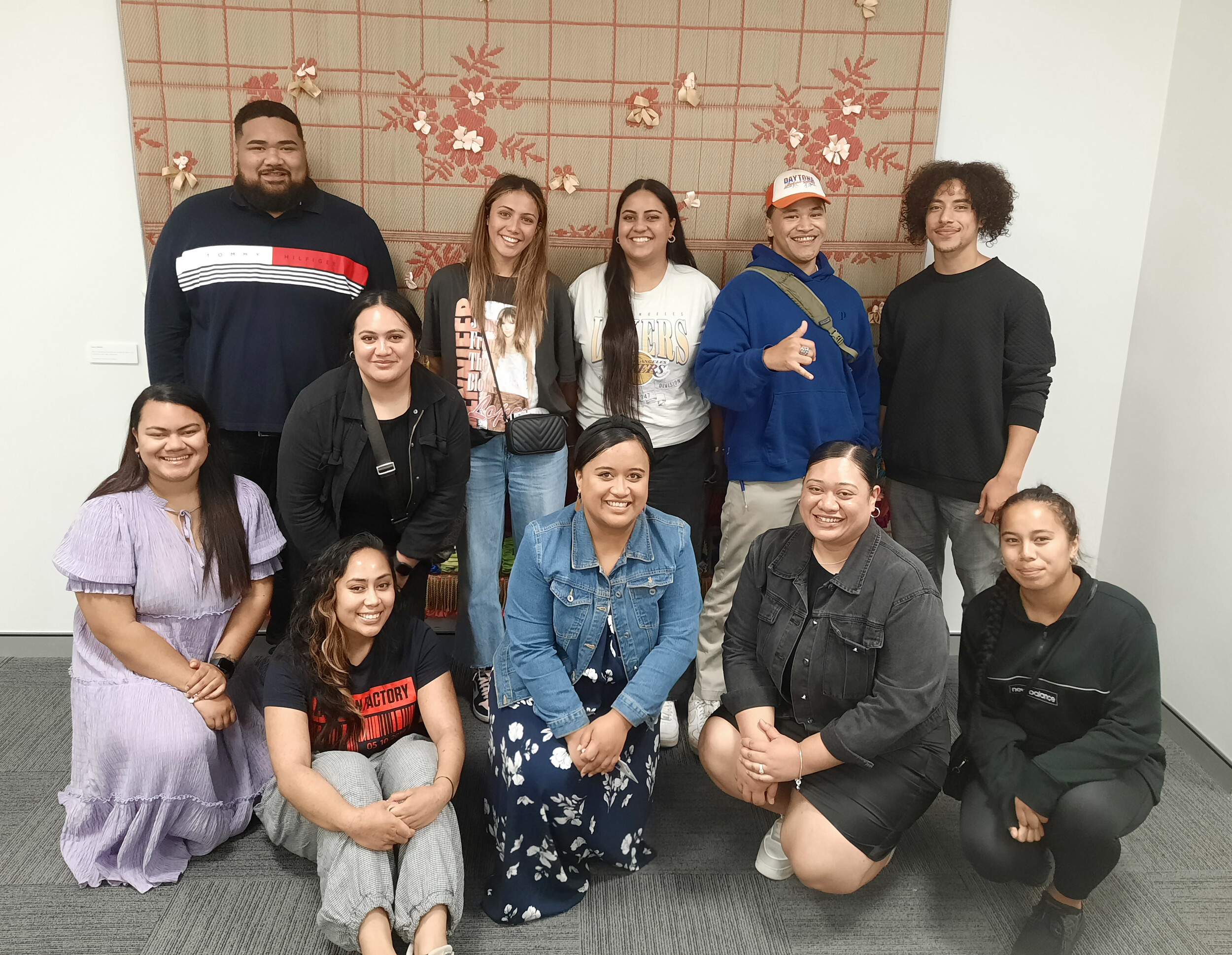 A group of young Pacific people smile for the camera in front of a floral woven mat.