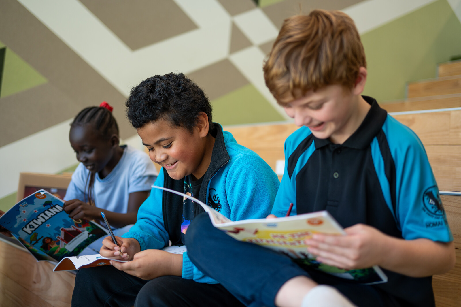 Three children in their school uniforms sit together on a bench smiling and writing in their workbooks 