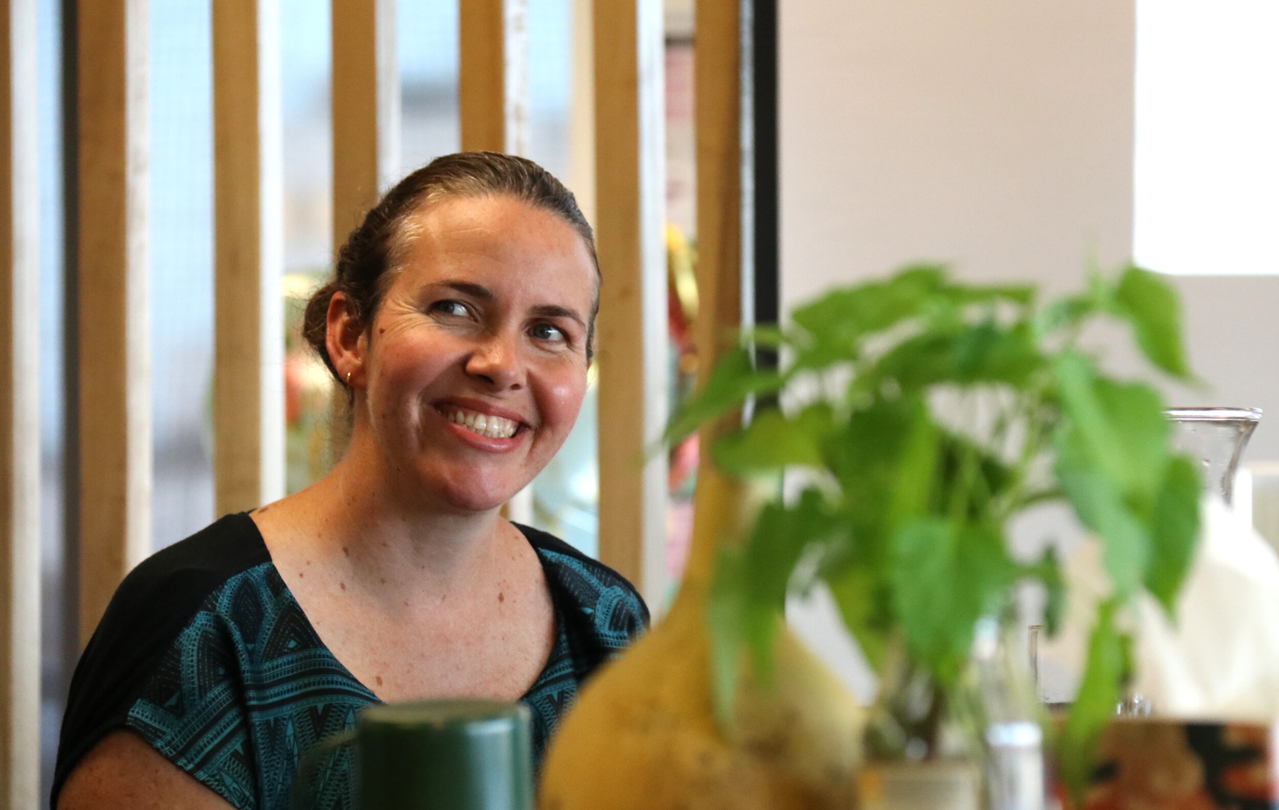 A head-and-shoulders image of a woman sitting inside smiling to someone off-camera, next to her is the top leaves of a green kumara plant and the top of a yellow dried gourd. 