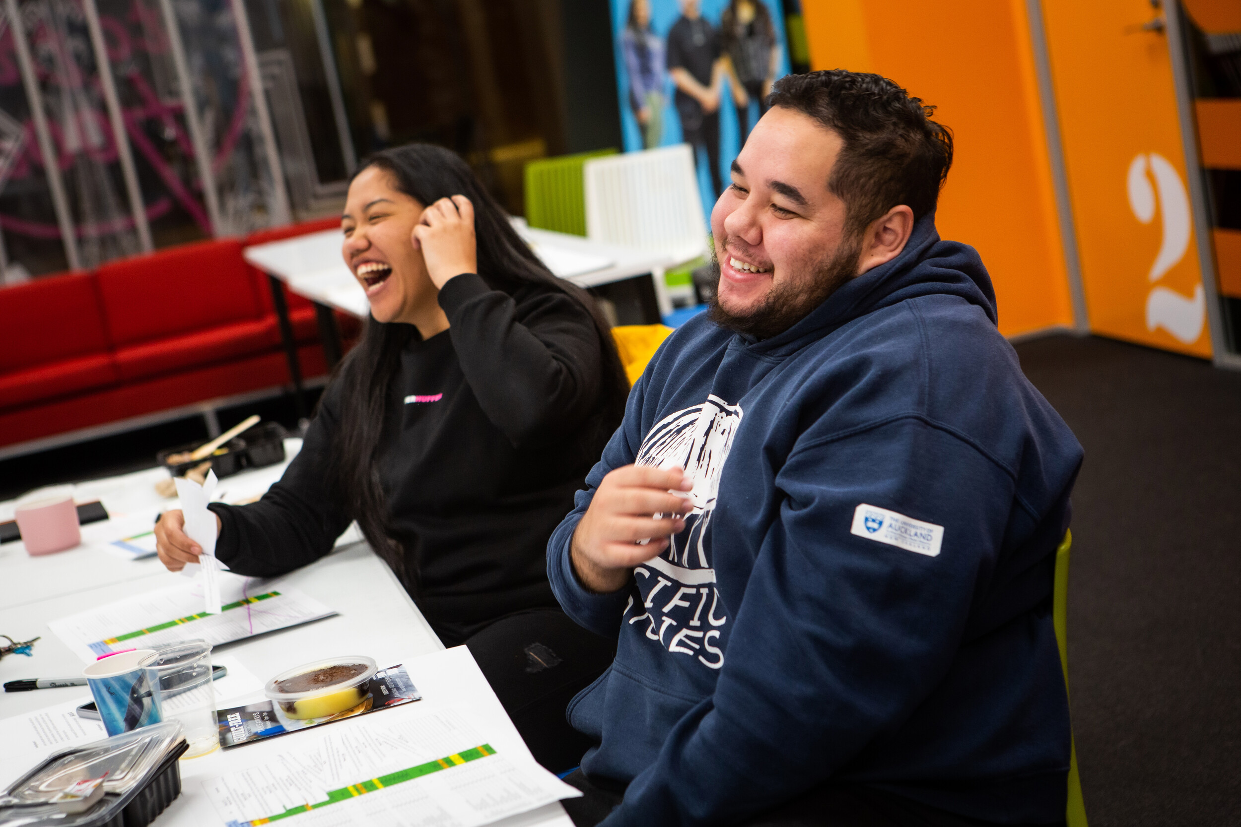 Two friends sitting at a table with some work sheets in front of them, smiling and laughing.